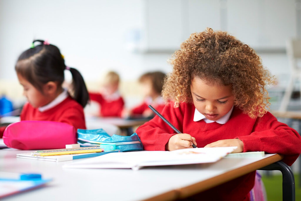 Schoolgirl in red uniform sitting at a desk in an infant school classroom drawing