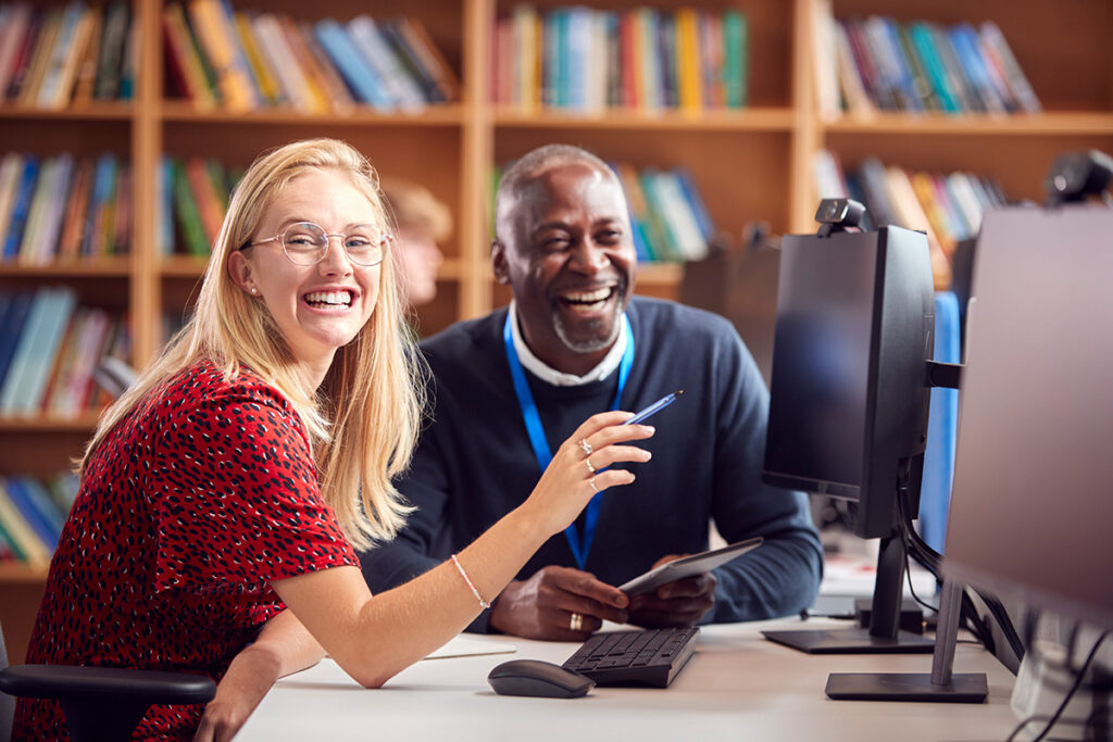 Two teacher colleagues sitting at a computer in a library laughing together