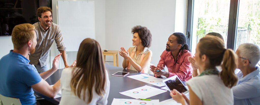 Group of teachers sitting around a table with paperwork while one teacher stands and speaks to the group